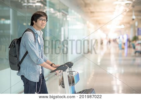 Young Asian man walking with airport trolley and his suitcase luggage in the international airport terminal arrival from travel abroad