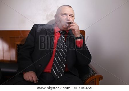 Alderen alvorlige business mand i jakkesæt ryge cigar på lædersofa i røget-up kontor. Fokusere på cigar