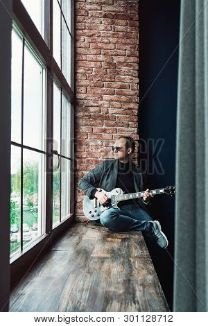 Man Singer Sitting On A Window Sill In A Headphones With A Guitar Recording A Track In A Home Studio