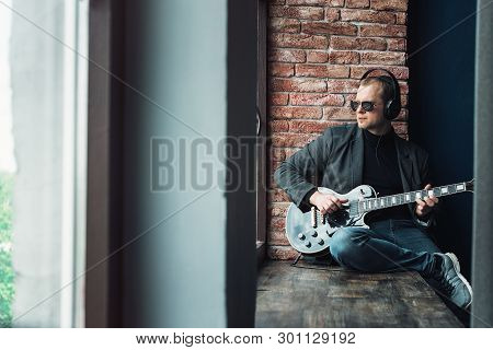 Man Singer Sitting On A Window Sill In A Headphones With A Guitar Recording A Track In A Home Studio