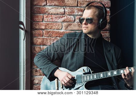 Man Singer Sitting On A Window Sill In A Headphones With A Guitar Recording A Track In A Home Studio