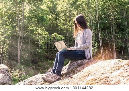 Young Beautiful Girl Is Working On A Laptop While Sitting On A Big Rock In The Forest. Freelance Con