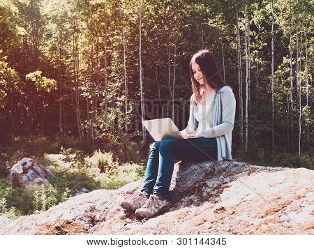 Young Beautiful Girl Is Working On A Laptop While Sitting On A Big Rock In The Forest. Freelance Con