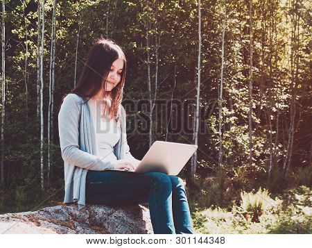 Young Beautiful Girl Is Working On A Laptop While Sitting On A Big Rock In The Forest. Freelance Con