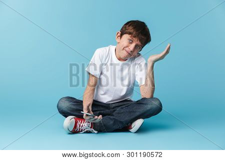 Image of european teen boy 10-12y with freckles wearing white casual t-shirt holding and using smartphone while sitting on floor isolated over blue background
