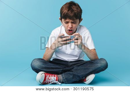 Image of caucasian brunette boy 10-12y with freckles wearing white casual t-shirt holding and using smartphone while sitting on floor isolated over blue background
