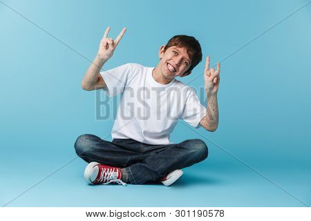 Image of cheery brunette boy 10-12y with freckles wearing white casual t-shirt smiling at camera while sitting on floor isolated over blue background