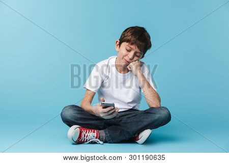 Image of european attractive boy 10-12y with freckles wearing white casual t-shirt holding and using smartphone while sitting on floor isolated over blue background