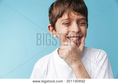 Image closeup of amusing caucasian boy 10-12y with freckles wearing white casual t-shirt smiling at camera isolated over blue background