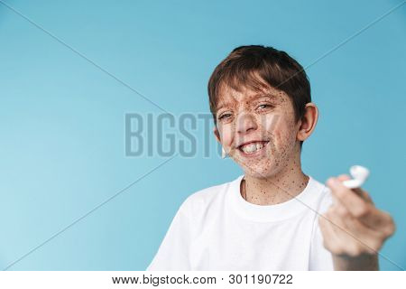 Image of attractive caucasian boy 10-12y with freckles wearing white casual t-shirt and earpods smiling at camera isolated over blue background