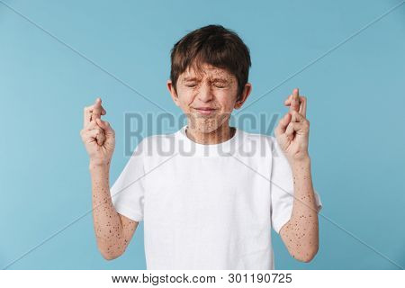 Image of caucasian boy 10-12y with freckles wearing white casual t-shirt keeping fingers crossed and begging for luck isolated over blue background