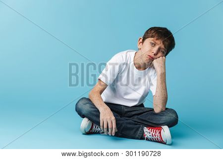 Image of european teen boy 10-12y with freckles wearing white casual t-shirt looking at camera while sitting on floor isolated over blue background