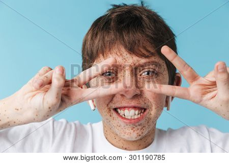 Image of content caucasian boy 10-12y with freckles wearing white casual t-shirt and earpods showing peace sign camera isolated over blue background