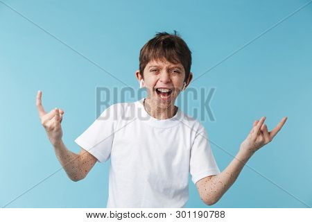 Image of ecstatic caucasian boy 10-12y with freckles wearing white casual t-shirt and earpods laughing at camera isolated over blue background