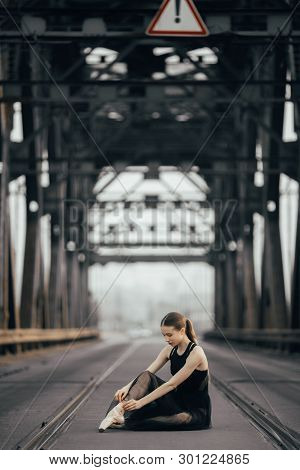 Ballerina Is Sitting And Tying Her Pointe Shoes Between Rails On The Road Against The Background Of 