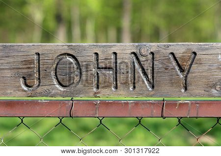 Dilapidated Vintage Retro Sign With Carved Johny On Top Mounted On Metal Pole Of Wire Fence With Gre