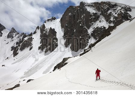 Snowy Mountains With Avalanche Traces, Sunlit Cloudy Sky, Hiker In Red With Ski Poles And Dog On Sno