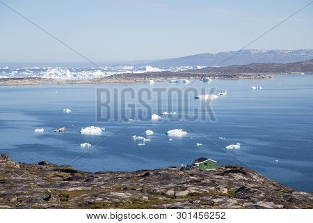 Ilulissat, Greenland - July 08, 2018: A Green Wooden House At The Coast With Icebergs In The Backgro