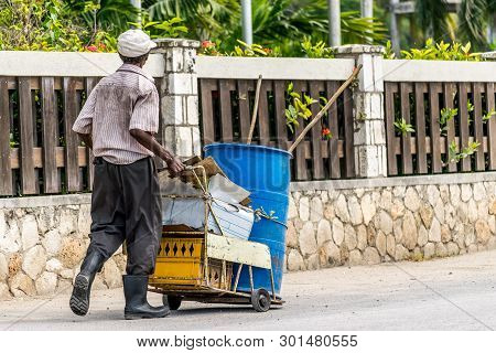 Black Elderly Man Wearing Rubber Boots And Dirty Clothing On The Street Pushes Cart With Plastic Dru