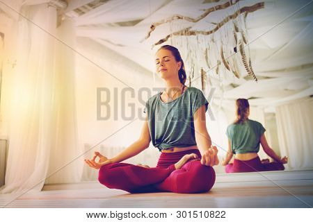 mindfulness, spirituality and healthy lifestyle concept - woman meditating in lotus pose at yoga studio with soft sunlight