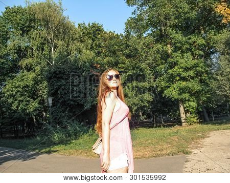 Beautiful Girl With Long Golden Hair In Sunglasses Looks At The Sun. Young Woman In Pink Clothes Wal