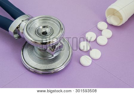 White Pills Falling Out Of A Plastic Tube Or Jar And The Head Of The Stethoscope On A Purple Backgro