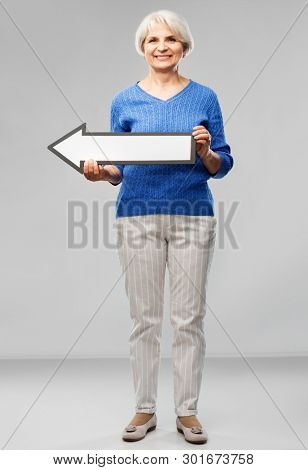 direction and old people concept - smiling senior woman in blue sweater holding big white leftwards thick arrow over grey background