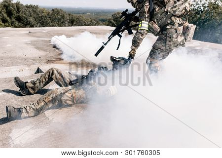 Armed Man In Camouflage With A Sniper Gun In His Hand, Pulling A Wounded Soldier
