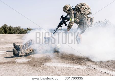 Armed Man In Camouflage With A Sniper Gun In His Hand, Pulling A Wounded Soldier In The Smoke