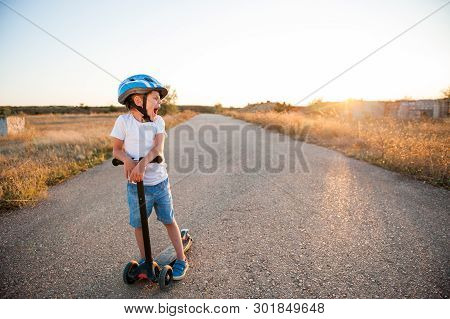 Sport Little Kid In Helmet And Blue Denim Shorts Yawn Standing On Empty Asphalt Road With Scooter On