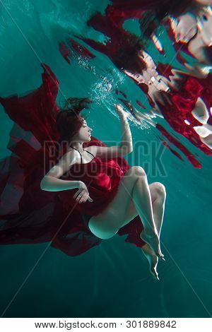 Mystical Underwater Portrait Of A Beautiful Young Woman In A Red Dress. The Girl Swims Under The Sur