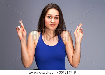 Body language and emotions concept. Glad dark haired beautiful satisfied girl beggs God to help her, keeps fingers crossed, wishes good luck, prays indoor isolated over gray studio background.