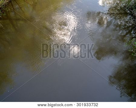 A Mirror Image In The Water Of The Blue Sky, Clouds On The Trees Surrounding The River. The Ripples 