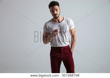 Serious looking young man holding his glasses and looking to the camera while wearing a white t-shirt and red pants, standing on gray studio background