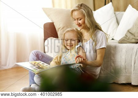 Happy Family. Blonde Young Mother Reading A Book To Her Cute Daughter While Sitting At Wooden Floor 
