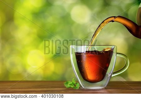 Black Tea With Mint Flavor Pouring From Glass Teapot Into Double Sided Wall Cup On Wooden Table With