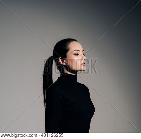 Portrait Of Young Woman In Black Turtleneck With Hairstyle Tail Against Of Grey Background. Side Vie