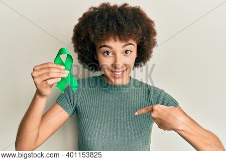 Young hispanic girl holding support green ribbon pointing finger to one self smiling happy and proud 