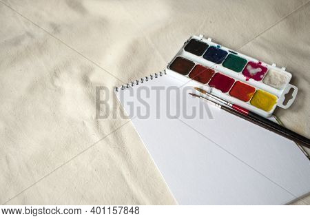 Set Of Watercolor Paints, Scetchbook And Paintbrush On A Beige Tablecloth With Copyspace.