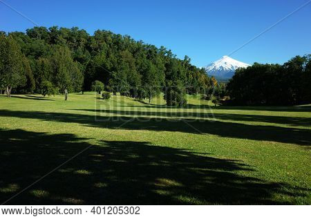 Nature of Chile. Beautiful landscape, sun and shadow, Green grass meadow and forest on hill, Snowy cone of Villarrica volcano under blue sky in sunny day. Green environment, Pucon