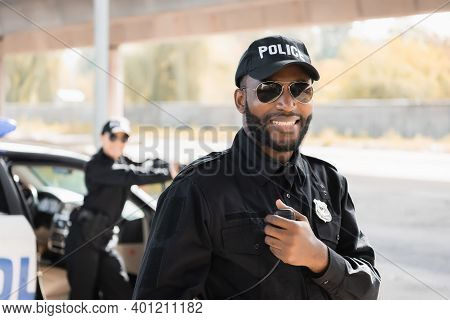 Happy African American Police Officer Looking At Camera While Holding Radio Set On Blurred Backgroun