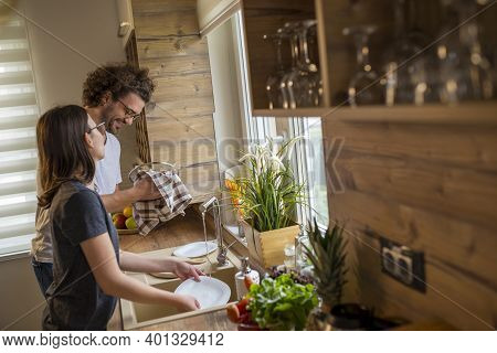 Young Couple In Love Doing The Washing Up And Drying Together After Lunch; Woman Washing While Man I