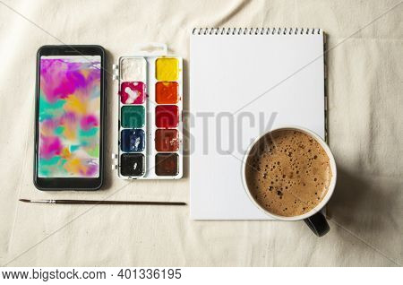 Set Of Watercolor Paints, Scetchbook, Paintbrush And A Cup Of Coffee For Breakfast.