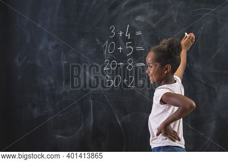 Beautiful Mixed Race Schoolgirl Standing In Front Of A Chalkboard, Smiling While Solving Math Equati