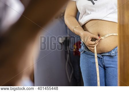 Mid Section Of An Indian Woman Measuring Fat Belly With Tape In Front Of Mirror
