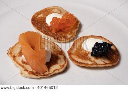 Mixed Blinis With Smoked Salmon, Rad And White Caviar Garnished With Sour Cream On A White Plate