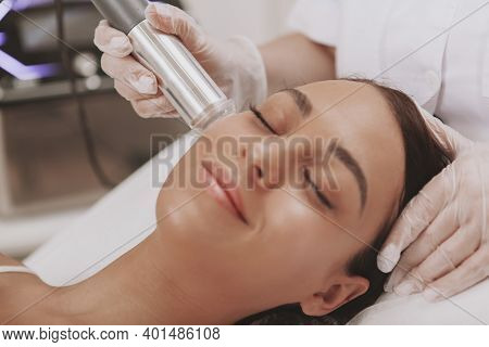 Charming Young Woman With Smooth Silky Skin Smiling, Receiving Microcurrent Facial Treatment By Prof