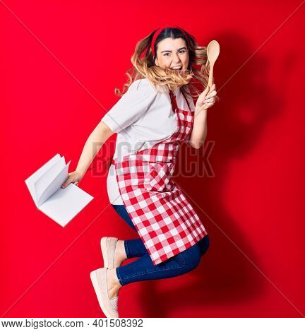 Young beautiful blonde cook woman wearing apron smiling happy. Jumping with smile on face holding recipe book and spoon over isolated red background