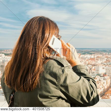 The Picture Shows A Back View Of A Caucasian Woman Talking On Her Smartphone From The Rooftop Of A B
