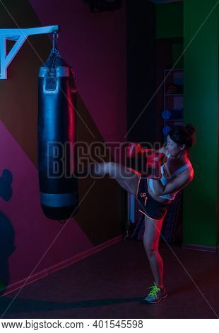 Female Boxer In Boxing Gloves Kicking A Punching Bag In Red Blue Neon Light Over Dark Background